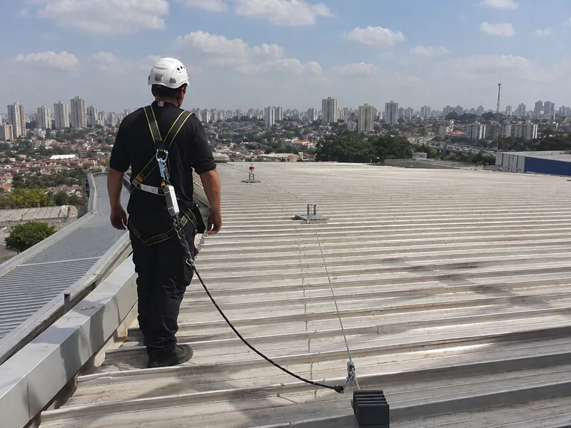 Técnico com capacete e cinto de segurança preso à linha de vida em cobertura industrial, com a cidade ao fundo