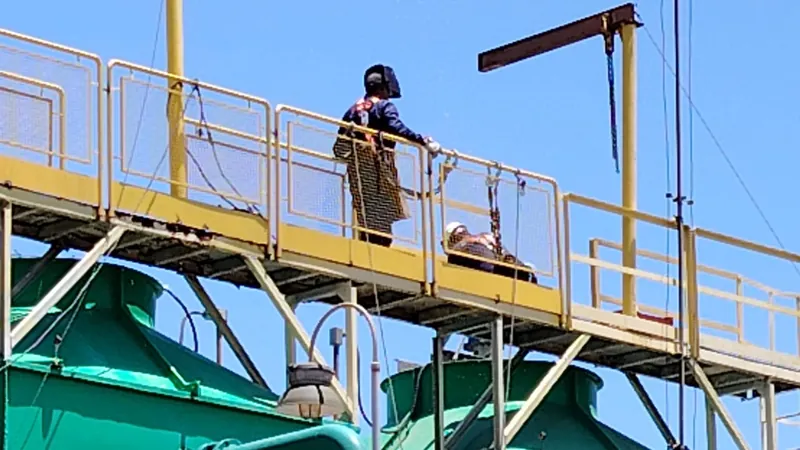 Técnico da VHE com capacete e cinto de segurança em plataforma elevada durante a inspeção, com céu azul ao fundo